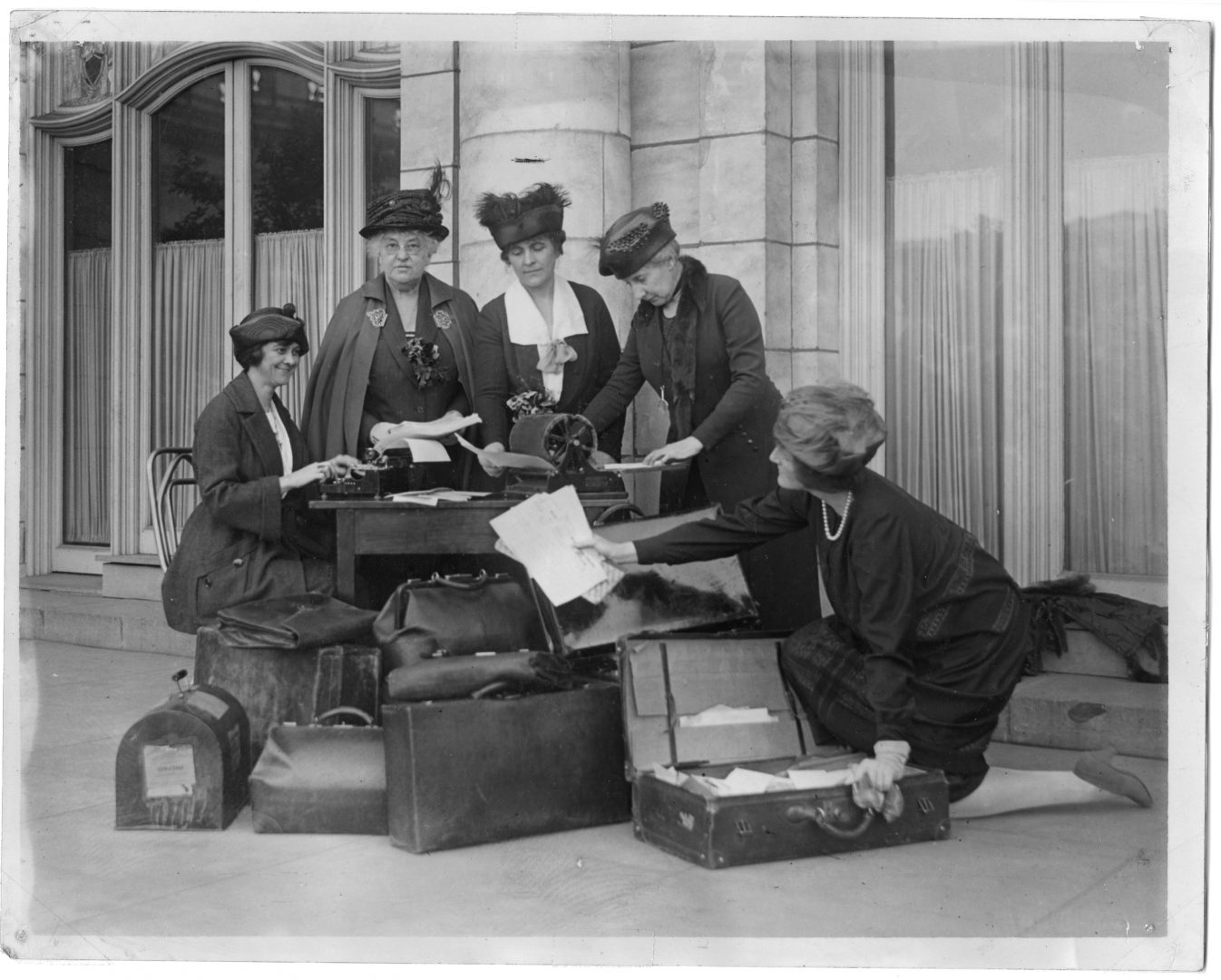 Five suffragists, including Marie Stuart Edwards (middle), a women's rights activist and reformer from Peru, Indiana, stand at a desk on the sidewalk with luggage during their western tour on behalf of the women's suffrage movement.